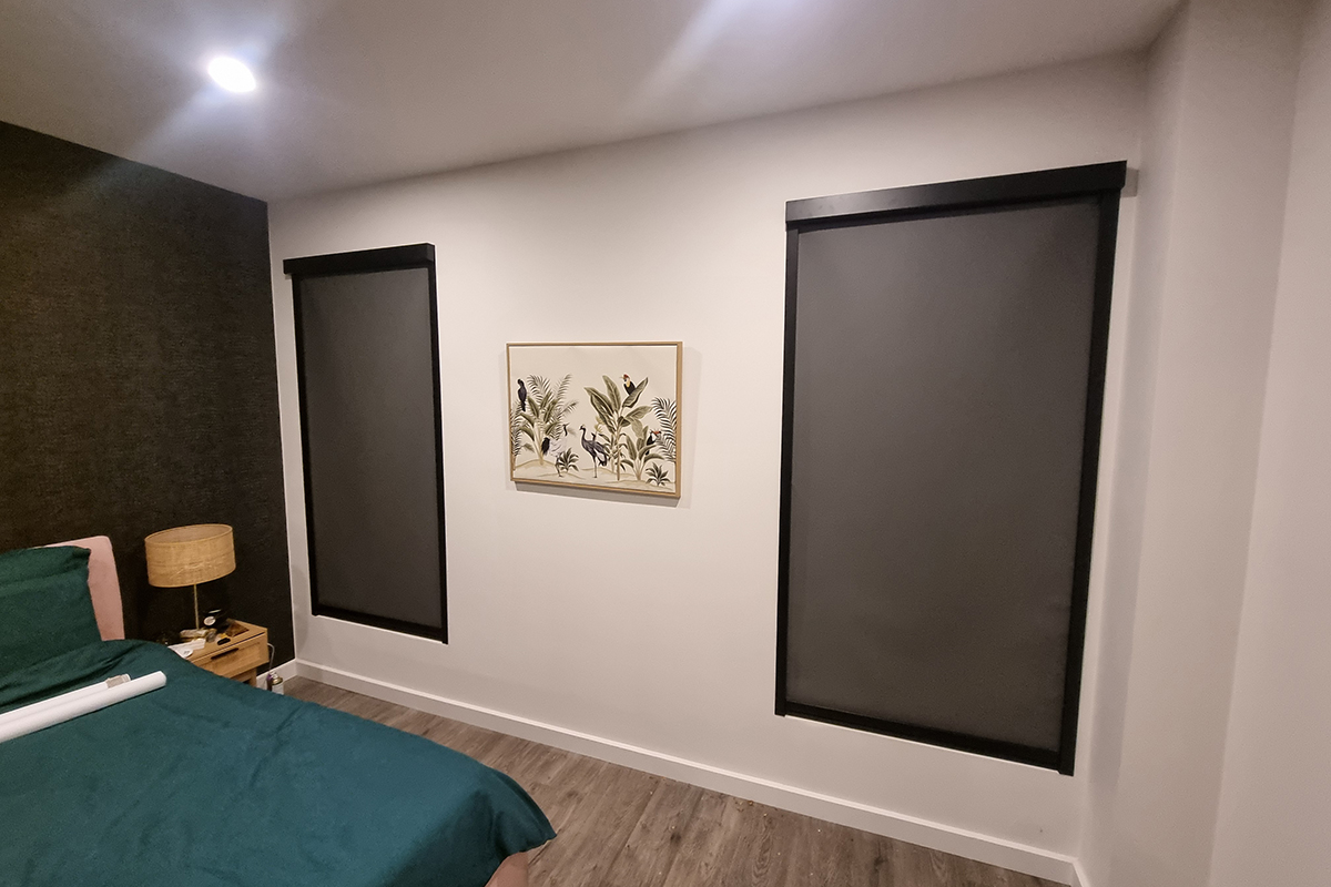 This image depicts a modern bedroom featuring two windows fitted with 100% TruBlockout blinds. These blinds are designed to provide complete darkness, making them ideal for improving sleep quality by blocking out all external light. The blinds have a sleek, minimalist design with black frames that blend seamlessly with the room's contemporary décor. The room itself is styled with a dark accent wall that complements the dark blinds, creating a cosy and restful atmosphere. A comfortable bed with dark green bedding sits in the corner, next to a small bedside table with a woven lamp. The neutral colour palette of the walls, along with the wooden floor, adds warmth to the space, while the botanical artwork between the windows introduces a touch of nature, enhancing the room's tranquil ambience. The overall design emphasises comfort and functionality, with the TruBlockout blinds playing a key role in ensuring a restful environment.