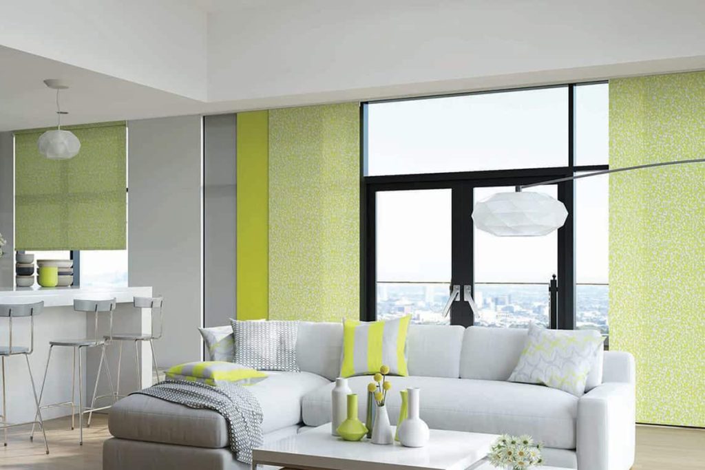 This image showcases a bright and modern open-plan living and kitchen area, featuring large windows dressed in stylish panel glide blinds in shades of yellow and grey. The blinds not only provide privacy and light control but also add a bold pop of colour and a contemporary touch to the space. The yellow panels bring energy and warmth, while the grey tones balance the vibrancy, creating a harmonious visual appeal. The kitchen area is sleek and minimalist, with glossy cabinets and a clean white countertop, complemented by modern bar stools. In the living area, a comfortable white sofa is adorned with cushions that echo the colour scheme of the blinds, tying the room's design elements together. The overall decor is fresh and inviting, with an emphasis on bright, cheerful colours and clean lines, making the space both functional and aesthetically pleasing.