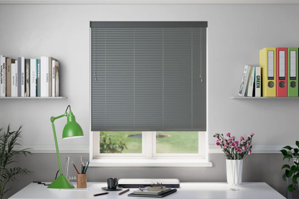 This image features a set of grey Venetian blinds installed over a window in a modern home office. The blinds are made of slimline aluminium horizontal slats, which can be adjusted to control the amount of light entering the room. Their purpose is to provide privacy, reduce glare on screens, and allow the user to easily adjust the light levels according to the time of day or activity. The blinds also contribute to the room's clean and organized aesthetic, complementing the minimalist decor and the functional workspace setup, which includes a desk, a green task lamp, and neatly arranged books and office supplies.