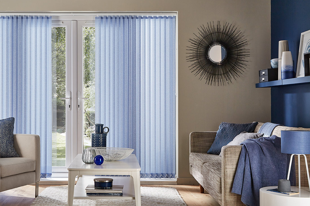 This image showcases a stylish and modern living room with large glass doors dressed in blue vertical blinds. The vertical blinds provide excellent control over light and privacy while adding a soft, calming hue to the space. The blinds’ vertical lines complement the room's clean and contemporary design, while the blue colour ties in with the overall colour scheme of the room. The room is furnished with a comfortable sofa and armchair, both adorned with blue and neutral-coloured cushions and throws, enhancing the cosy atmosphere. A sleek white coffee table sits in the centre, decorated with minimalist accessories that add a touch of elegance. The decor is completed with a sunburst mirror on the wall and various vases and decorative items that reinforce the room's serene and cohesive aesthetic. Overall, the vertical blinds serve both functional and aesthetic purposes, enhancing the room's light control while contributing to its modern and inviting style.